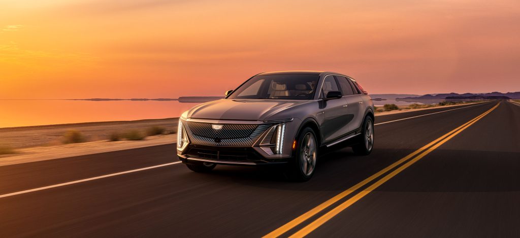 A silver Cadillac LYRIQ driving on a highway next to a calm body of water at sunset.