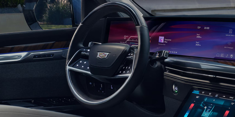 Driver's side interior perspective of a Cadillac vehicle, showing the steering wheel with the brand emblem, a massive sweeping digital dashboard display, and a lower center console screen for climate control.
